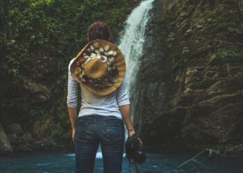 Frau mit Strohhut und Kamera vor einem Wasserfall