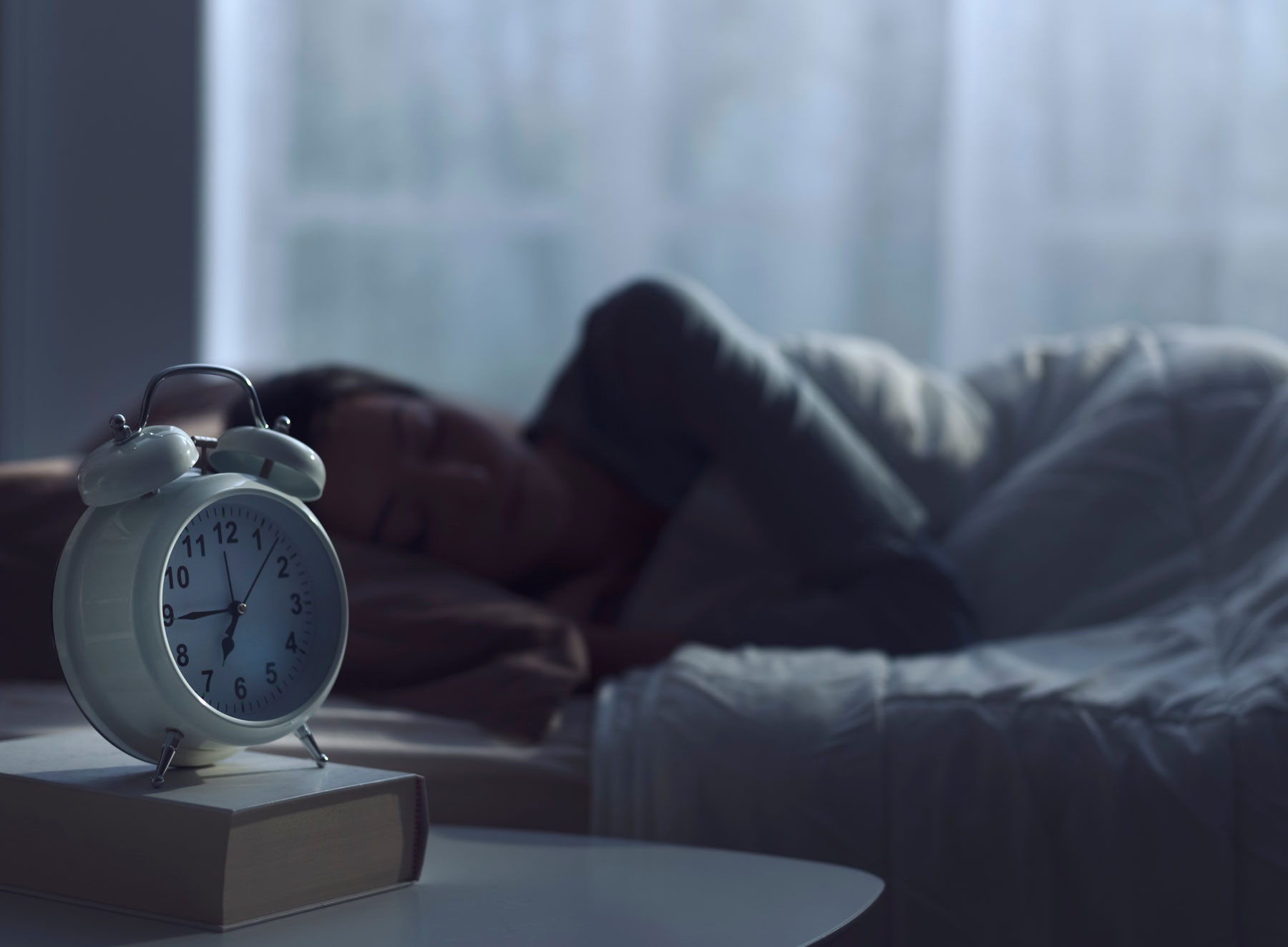 Ein klassischer Wecker auf dem Nachttisch, dahinter schlafend eine Frau in ihrem Bett